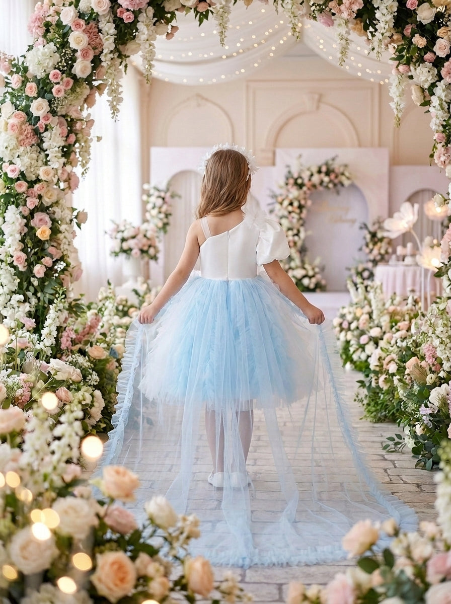Back view of a child wearing a white one-shoulder dress with a blue-and-white ruffled skirt and long sheer train.