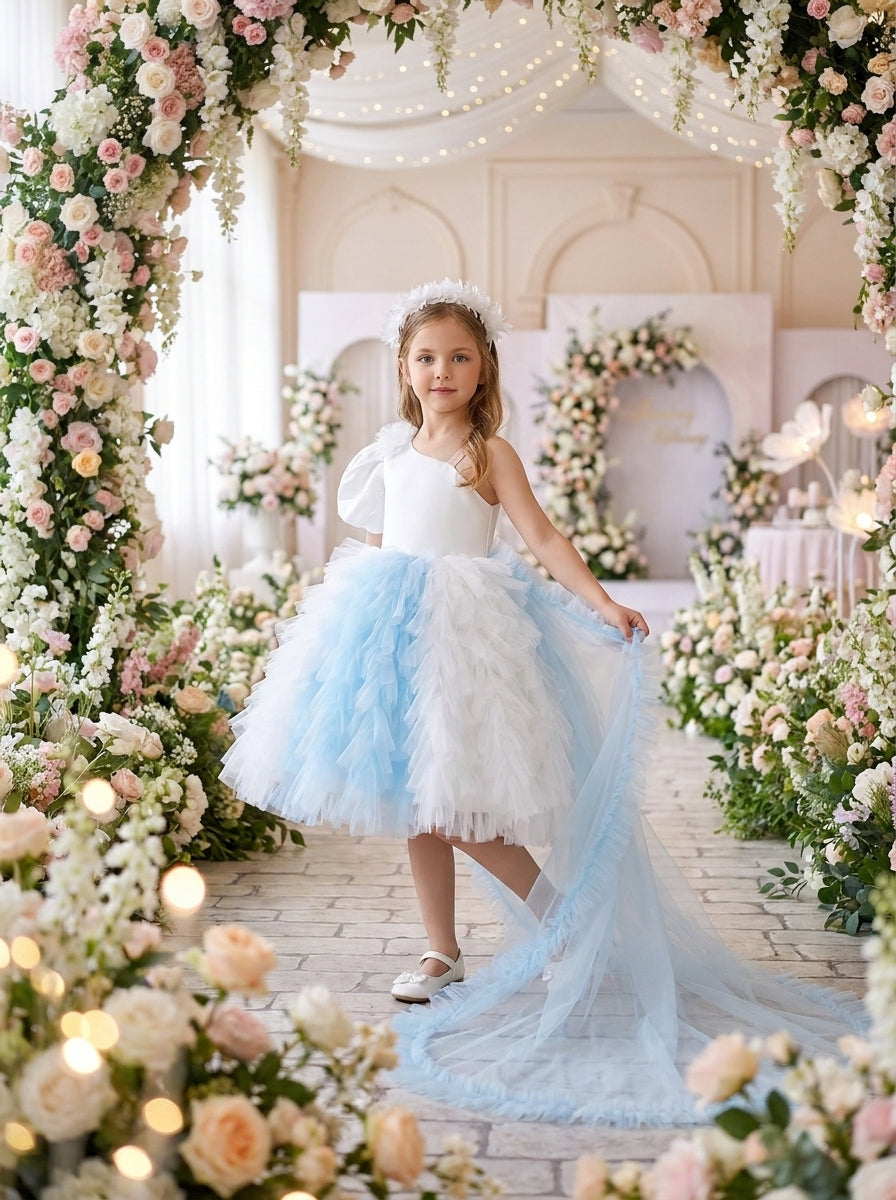 Front view of a child wearing a white one-shoulder dress with a blue-and-white ruffled skirt and long sheer train.