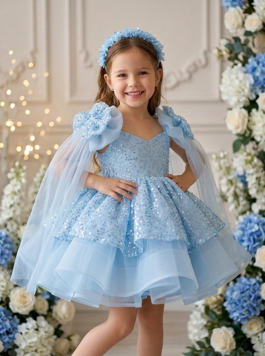 Front view of a child wearing a light blue sequinned dress with shoulder rosettes, sheer cape sleeves and a layered ruffle skirt.