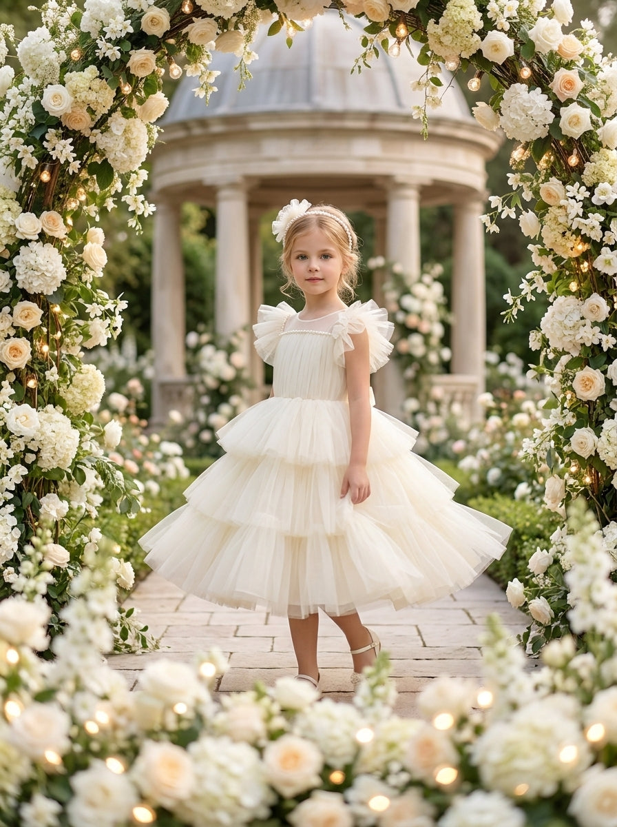 Front view of a child wearing a cream dress with ruffled shoulders, a pleated bodice and tiered skirt.