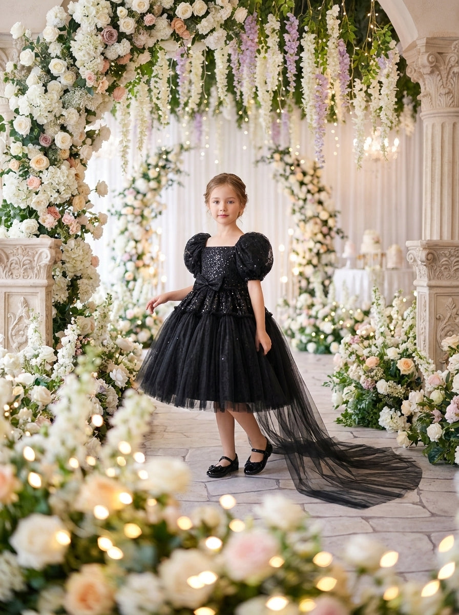 Front view of a child wearing a black dress with shiny puff sleeves, a waist bow and long sheer train.