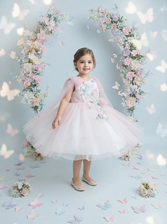 Front view of a child wearing a pale pink dress with pastel flowers, butterfly decorations and shoulder drapes.